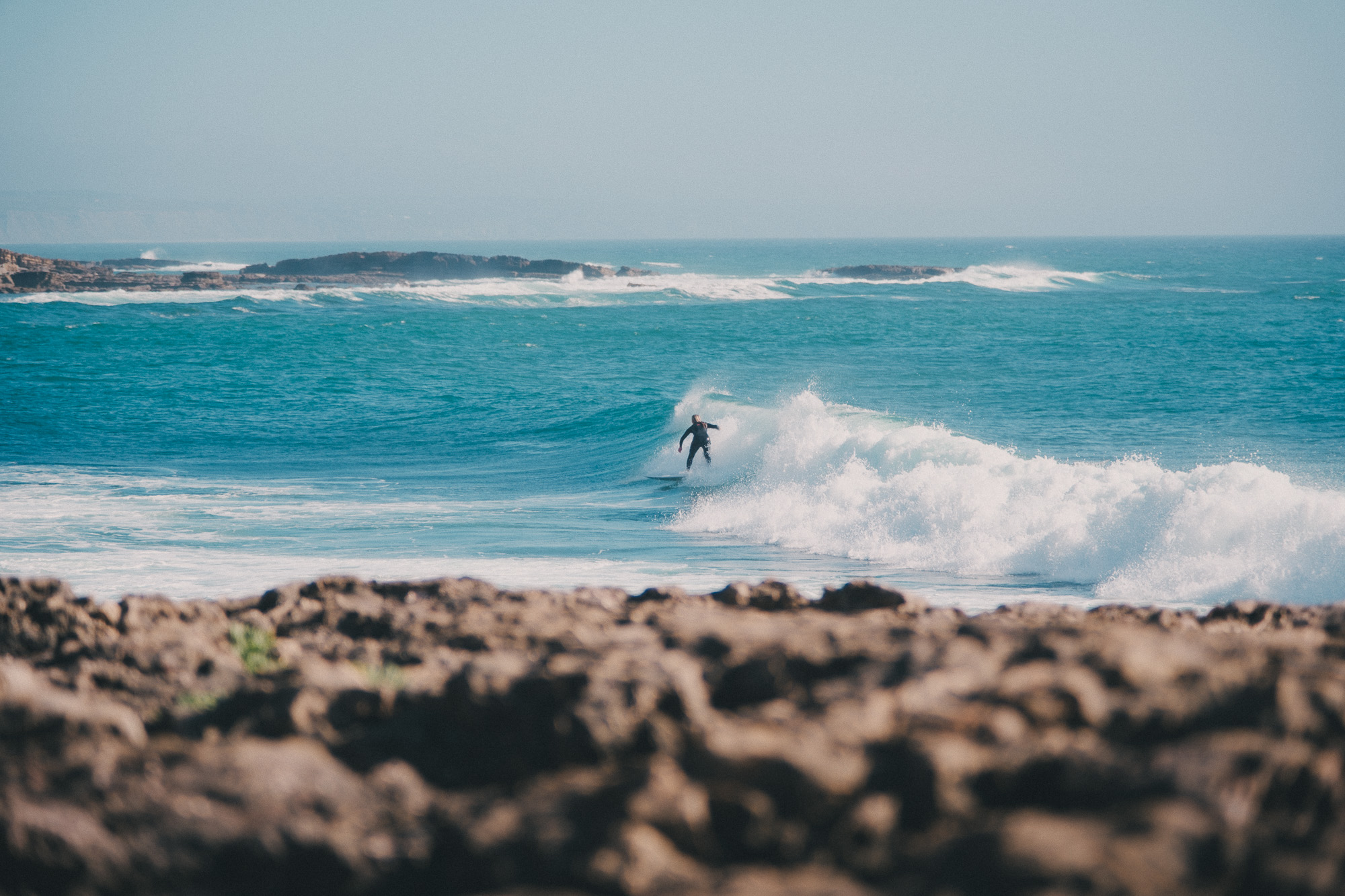 Minimalist photography of a lone surfer on a large ocean wave in Ericeira, Portugal. High contrast water textures and white sea foam against a deep blue Atlantic background.